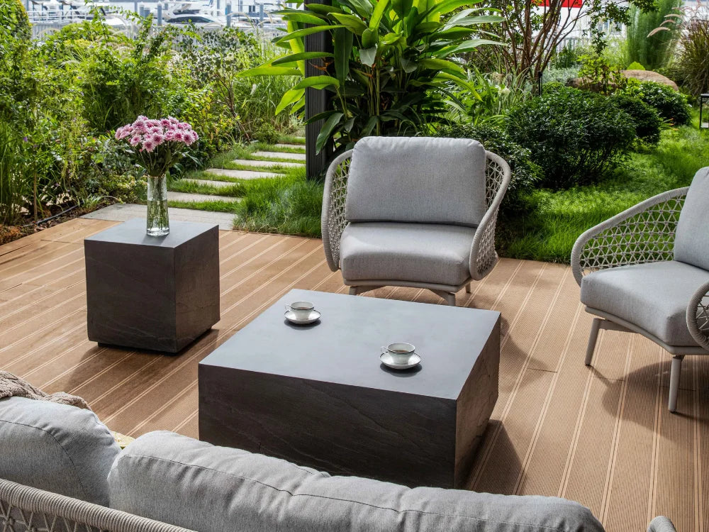 Modern outdoor patio furniture set including a dark concrete coffee table, two gray cushioned wicker armchairs, and a matching side table on a wooden deck with a lush green garden background.