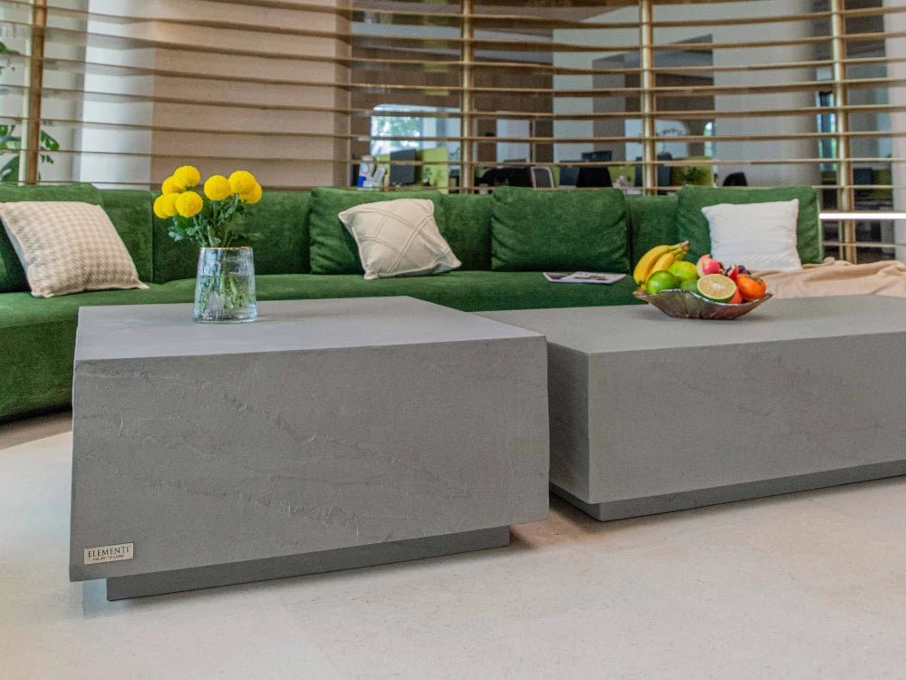Modern concrete coffee tables in a bright, contemporary living space with a green sectional sofa, decorative pillows, and fruit and flowers on top.