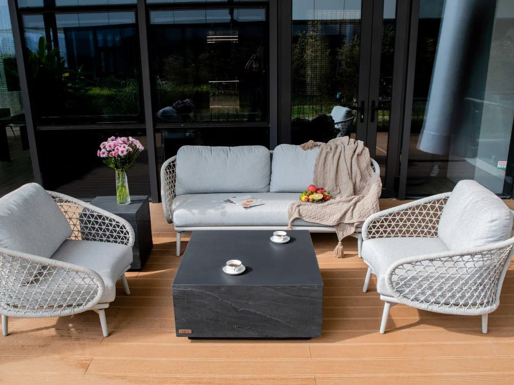 Modern outdoor patio furniture set featuring a light gray fabric sofa, two matching armchairs with woven white frames, a dark gray square coffee table, and a small side table with pink flowers, set on a wooden deck in front of a glass-walled building.