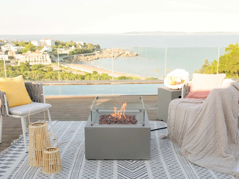 A modern square fire pit table with a glass top and visible flames, placed on a patterned rug on a spacious outdoor terrace overlooking a coastal bay and beach.