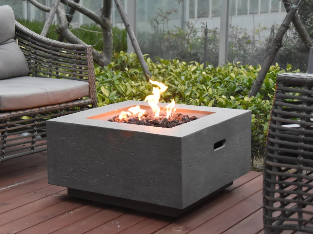 A square concrete outdoor fire pit table with a lit fire inside, placed on a wooden deck between wicker patio chairs with cushions.
