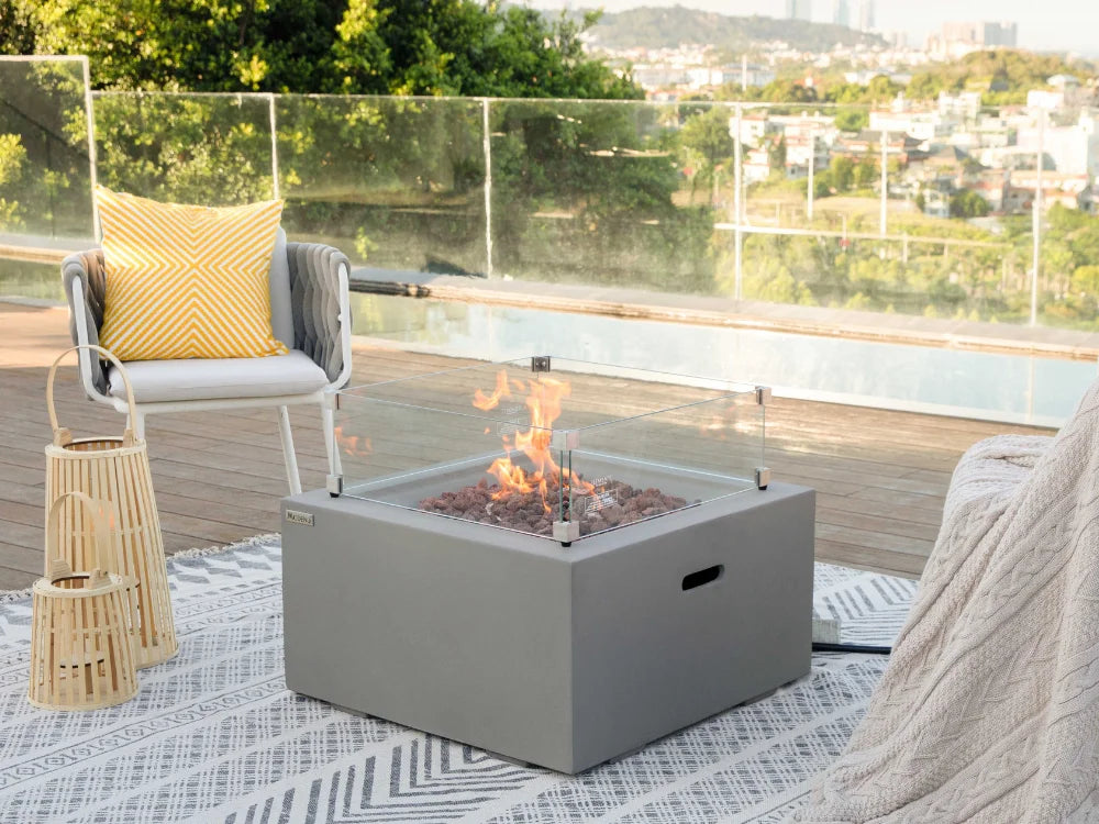 A square outdoor fire pit table with a gray metal body and glass wind guard, set on a patio next to a chair with a yellow pillow. The fire pit contains glowing fire glass and is lit with visible flames, with a scenic pool and city view in the background.