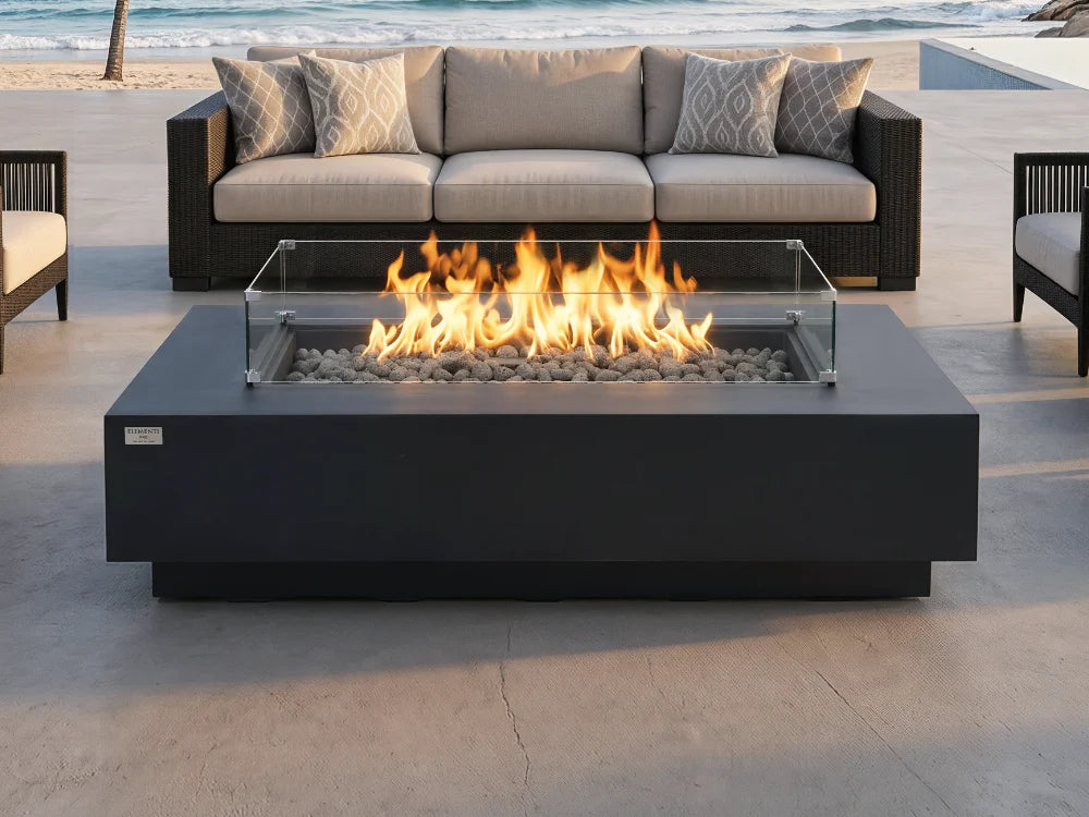 Elementi rectangular dark gray outdoor fire pit table with a glass wind guard and active flames, positioned in front of a modern outdoor sofa on a patio with a beach background.