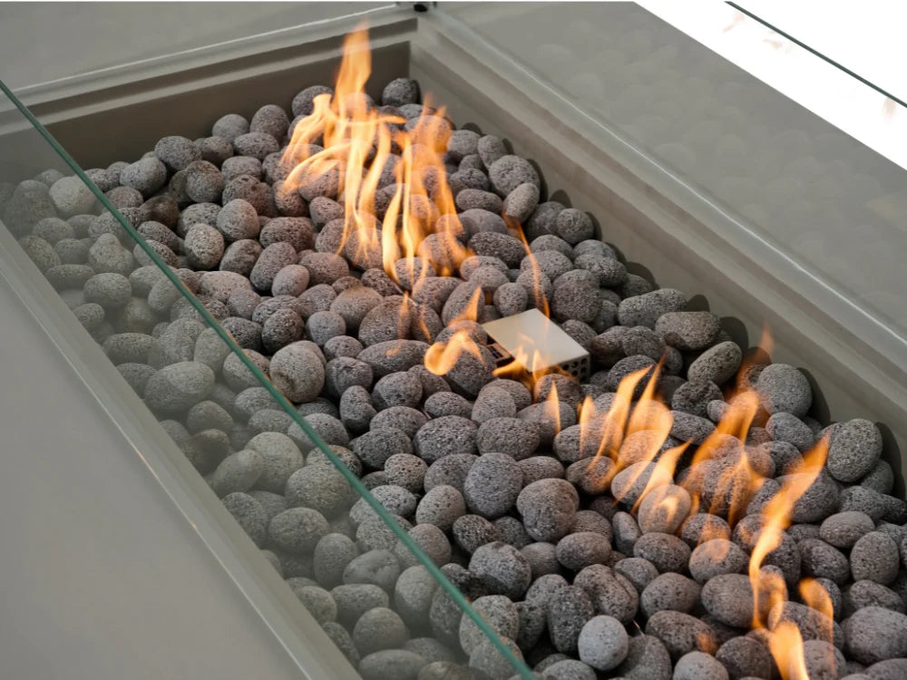 A close-up view of Elementi rectangular fire pit with gray pebbles and active orange flames inside a modern light gray metal frame with a clear glass wind guard.