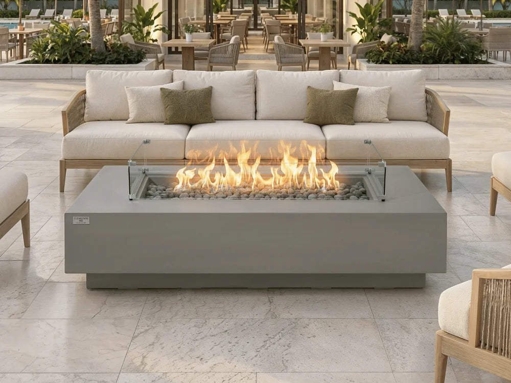 Elementi rectangular outdoor fire pit table with a glass wind guard and visible flames, placed in front of a light beige outdoor sofa on a tiled patio.