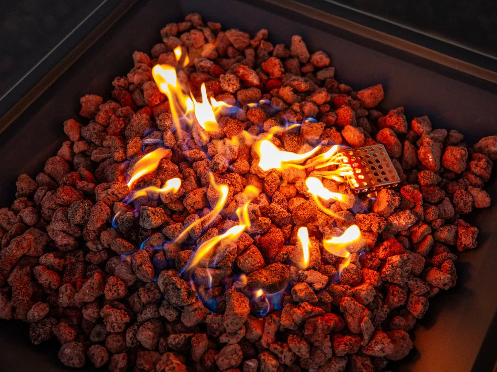 Close-up of a Elementi fire pit filled with glowing red and orange stones and bright yellow-orange flames, with a small metal grate visible near the center-right.