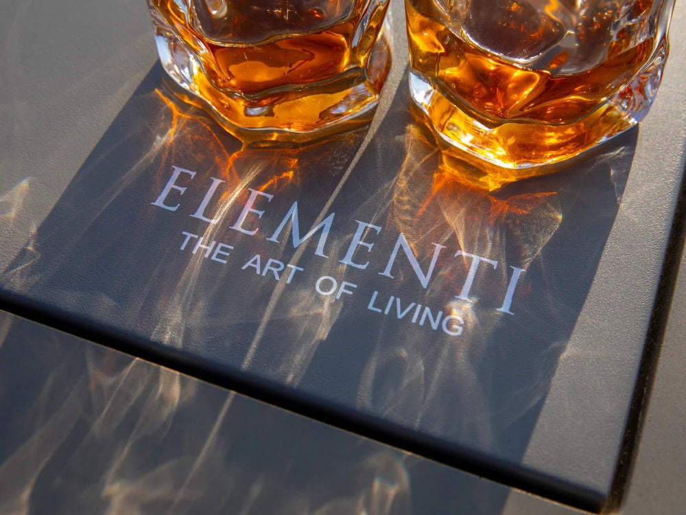 Two faceted glass whiskey tumblers filled with amber liquor sit on a dark tray printed with the brand name ELEMENTI and the phrase 'THE ART OF LIVING'.