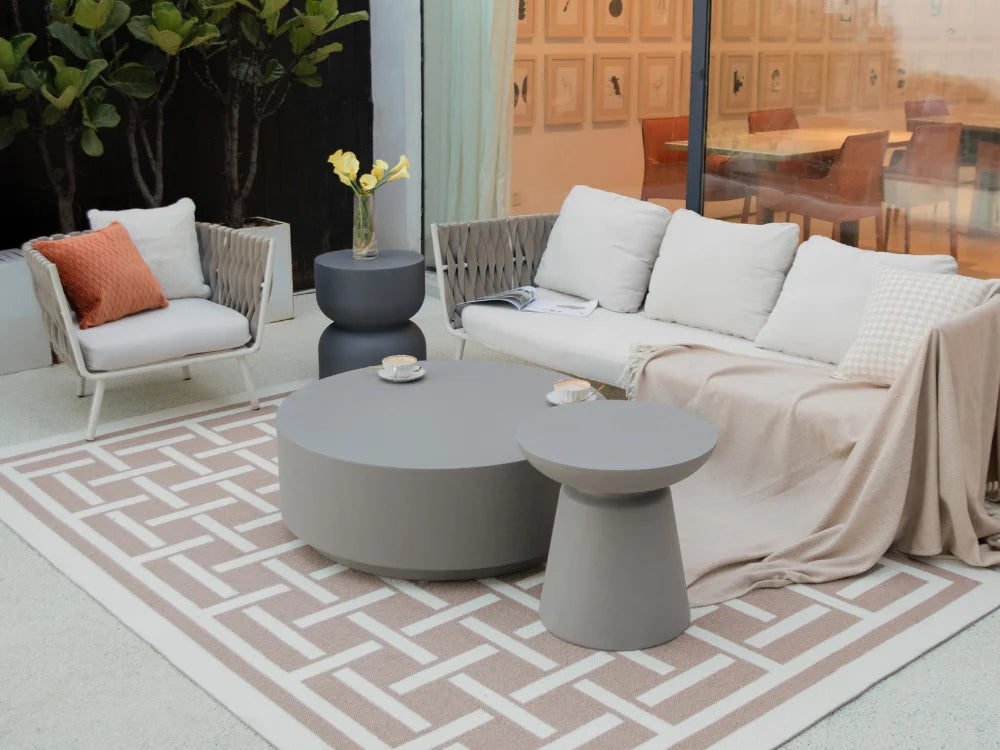 Modern outdoor patio furniture set including a gray round coffee table, matching side table, woven armchair with white cushions, and a sofa draped with a beige throw blanket, arranged on a geometric rug.