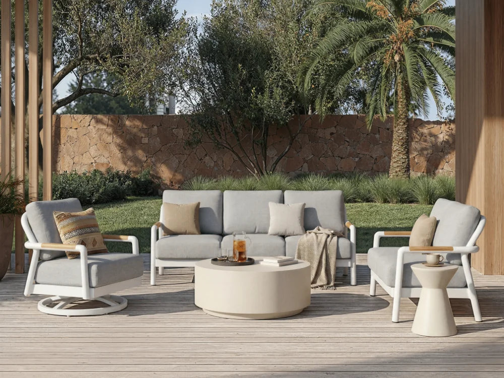Elementi modern outdoor patio furniture set including a three-seat sofa, two armchairs, a round coffee table, and a small side table on a wooden deck with a stone wall and palm tree in the background.