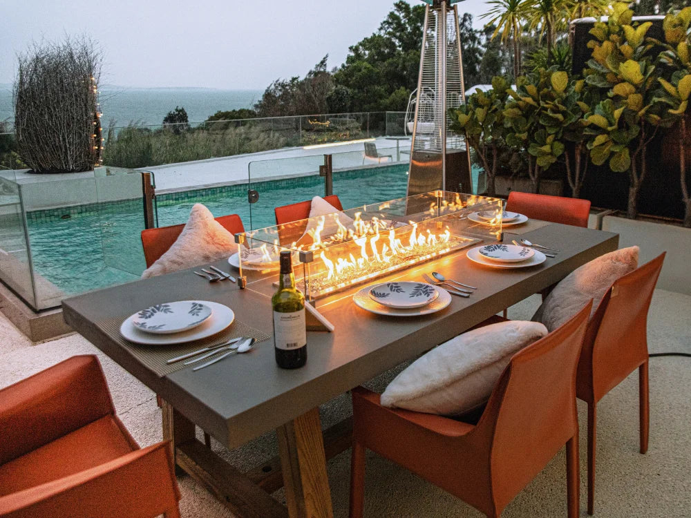 Elementi outdoor dining fire table on a patio with orange-brown chairs, plates, cutlery, a wine bottle, and a glowing linear fire pit. A glass railing overlooks a pool and ocean view, with plants and a tall patio heater in the background.