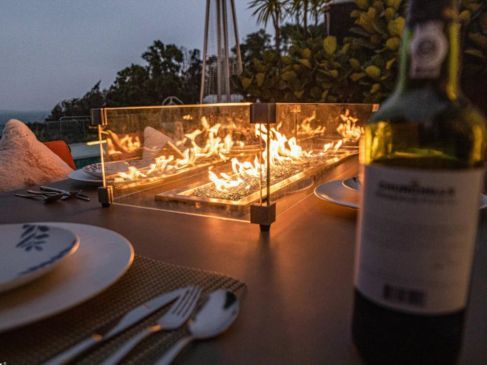 Outdoor patio dining scene at dusk with a rectangular glass fire pit centered on a table, glowing flames, plates, utensils, and a wine bottle in the foreground.