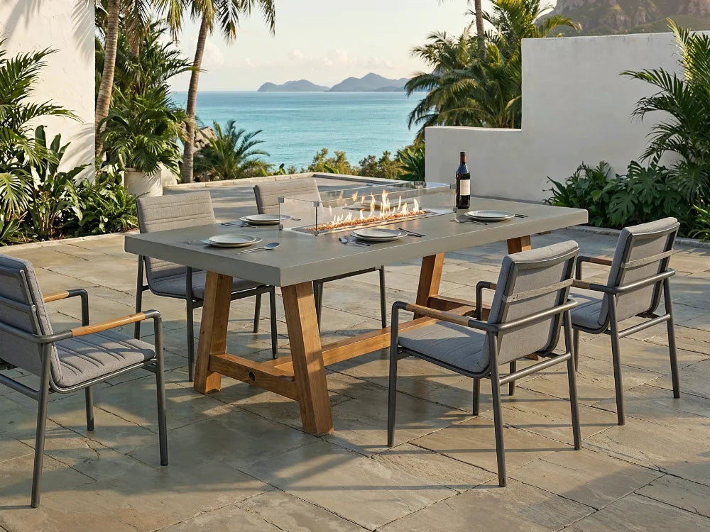 Outdoor dining set on a stone patio overlooking the ocean, featuring a rectangular concrete-top table with a built-in fire pit and six gray cushioned metal chairs.